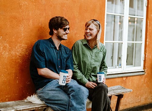 man and woman sitting together on a bench against an ochre wall, each holding a REUSEABLE cup