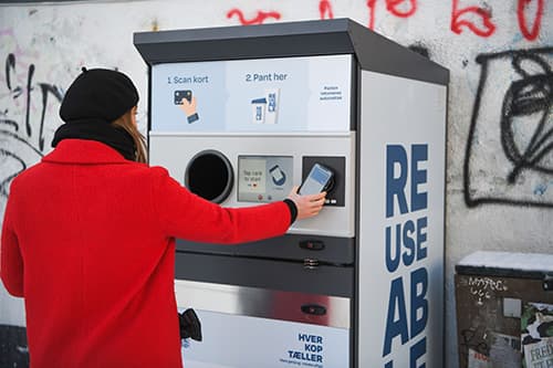 Woman in red coat and black hat tapping phone on TOMRA Reuse machine
