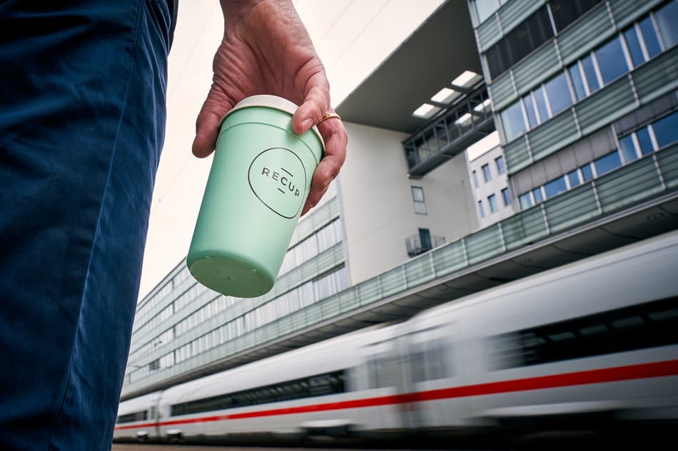 Man holding a RECUP in right hand with train in background